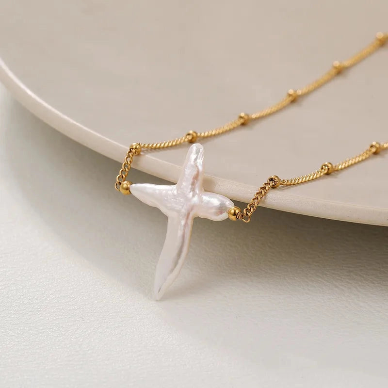 The Celestial Pearl Cross Necklace featuring an organic white Baroque pearl pendant on a gold titanium steel chain on a light background
